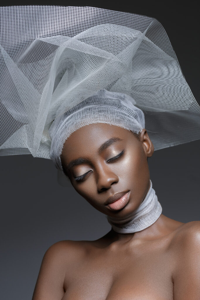 Beautiful young black woman with natural make up and glowing skin in white veil fashion hat. Beauty shot on grey background. Copy space.